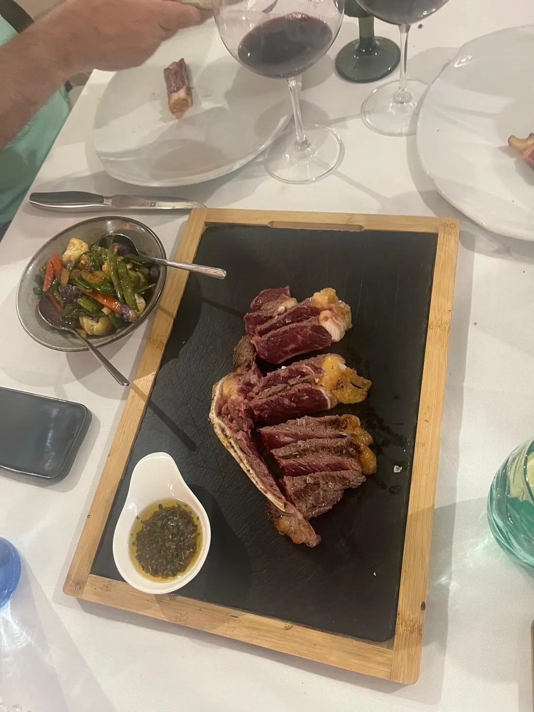 Chuletón fileteado sobre una pizarra, con chimichurri y verduras, en la mesa con copas de vino